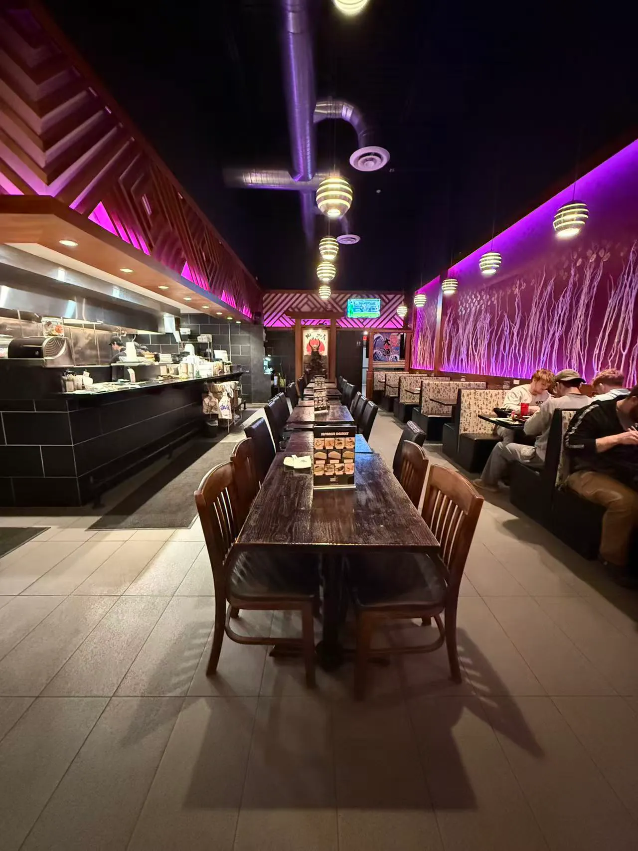 A dimly lit sushi restaurant interior with purple accent lighting, wooden tables, booth seating, and an open kitchen visible on the left.
